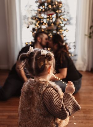 A small child watches a Christmas tree with twinkling lights while a couple embraces in the background.
