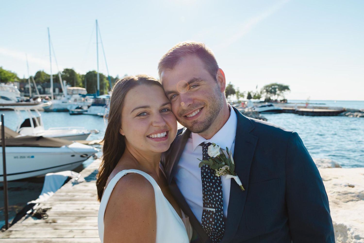 A romantic wedding couple poses on a sunny dock with boats and a marina in the background.