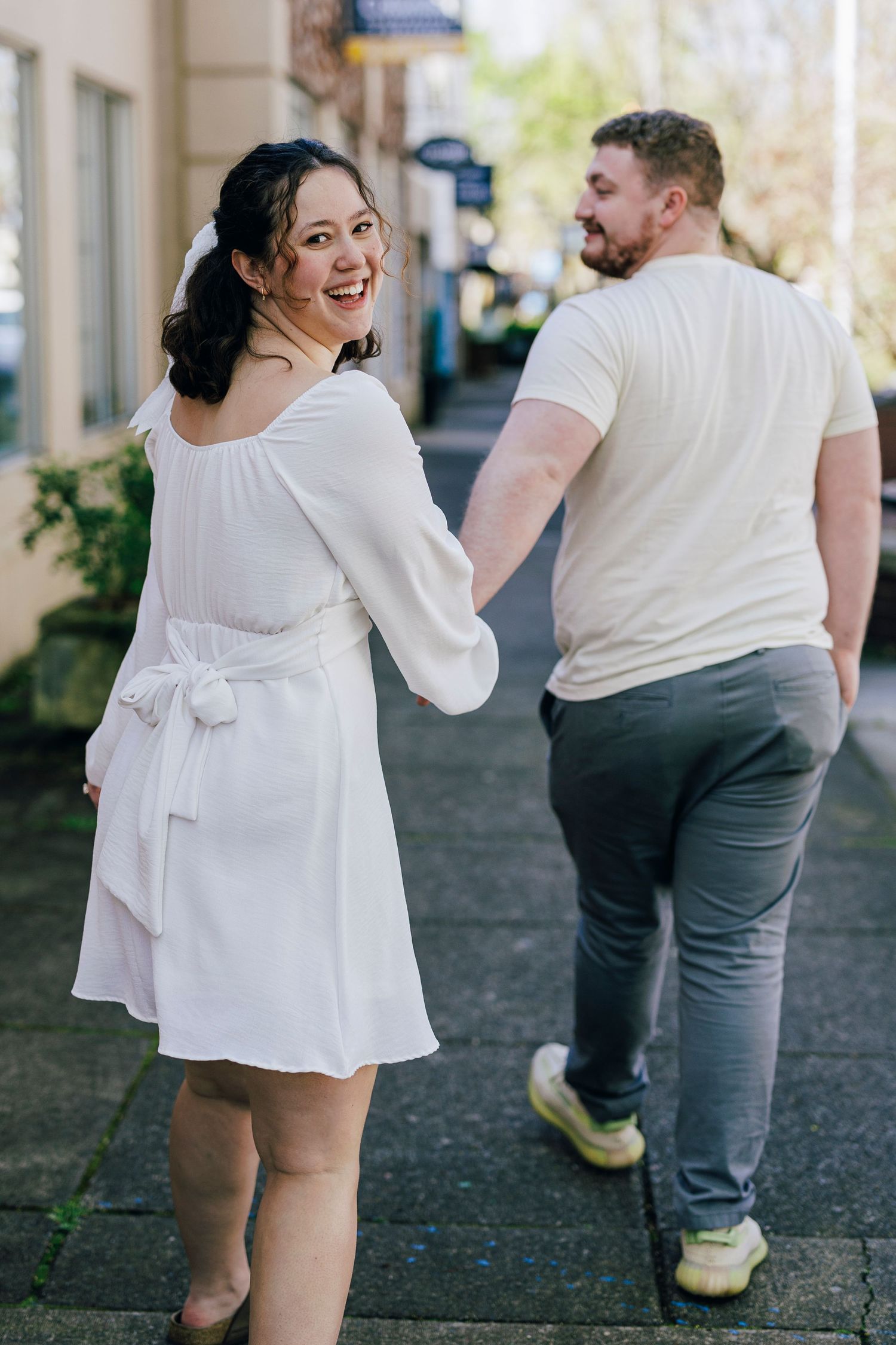 A couple in white outfits walking together along a charming urban sidewalk with storefronts in the background.