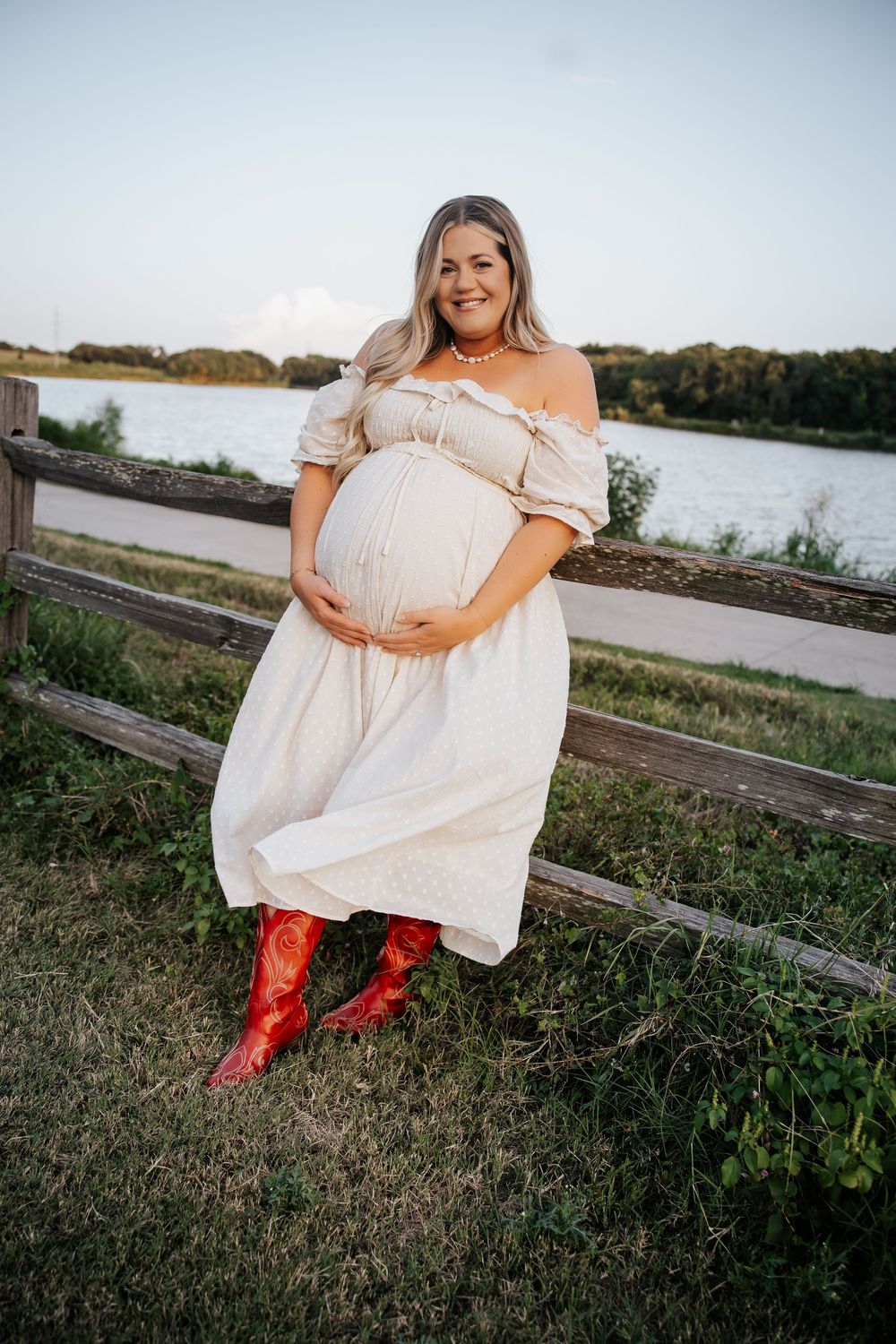 Smiling pregnant woman in white off-shoulder dress and red cowboy boots holding belly near a wooden fence.