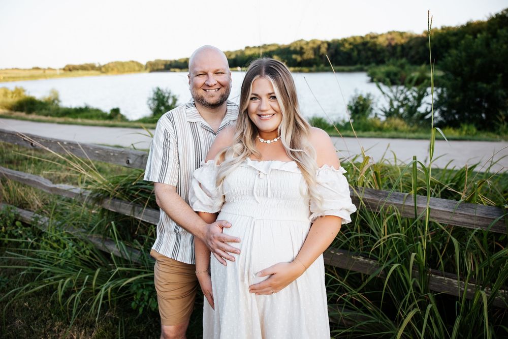 Smiling expectant couple standing by a wooden fence near a lake, woman in white off-shoulder maternity dress.
