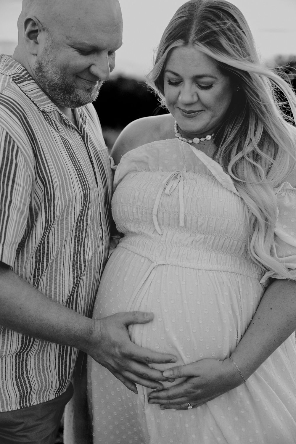 Black and white maternity photo of couple tenderly looking down at pregnant belly in outdoor setting.