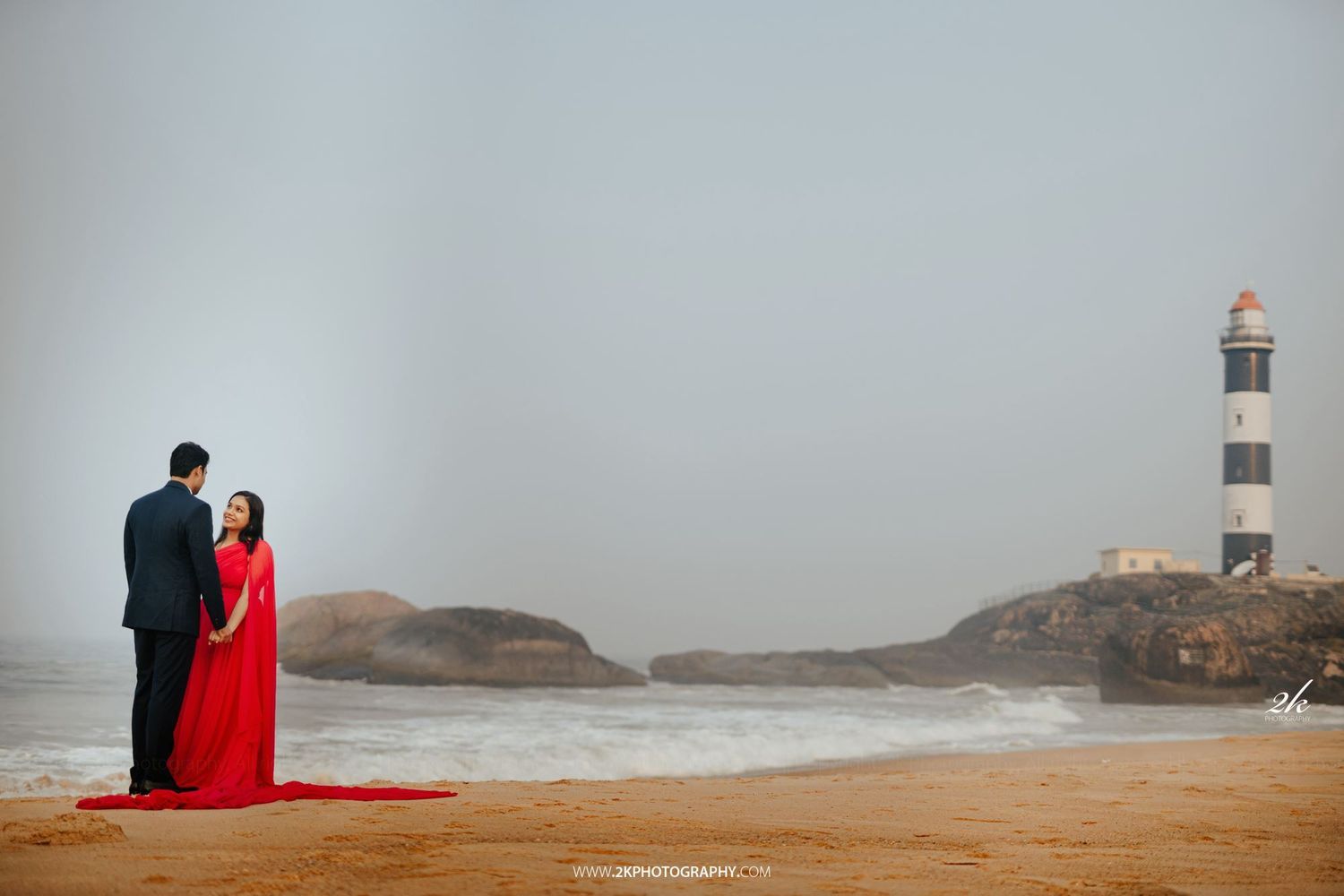 A couple stands on a misty beach near a lighthouse, with the woman wearing a flowing red dress and man in formal attire.