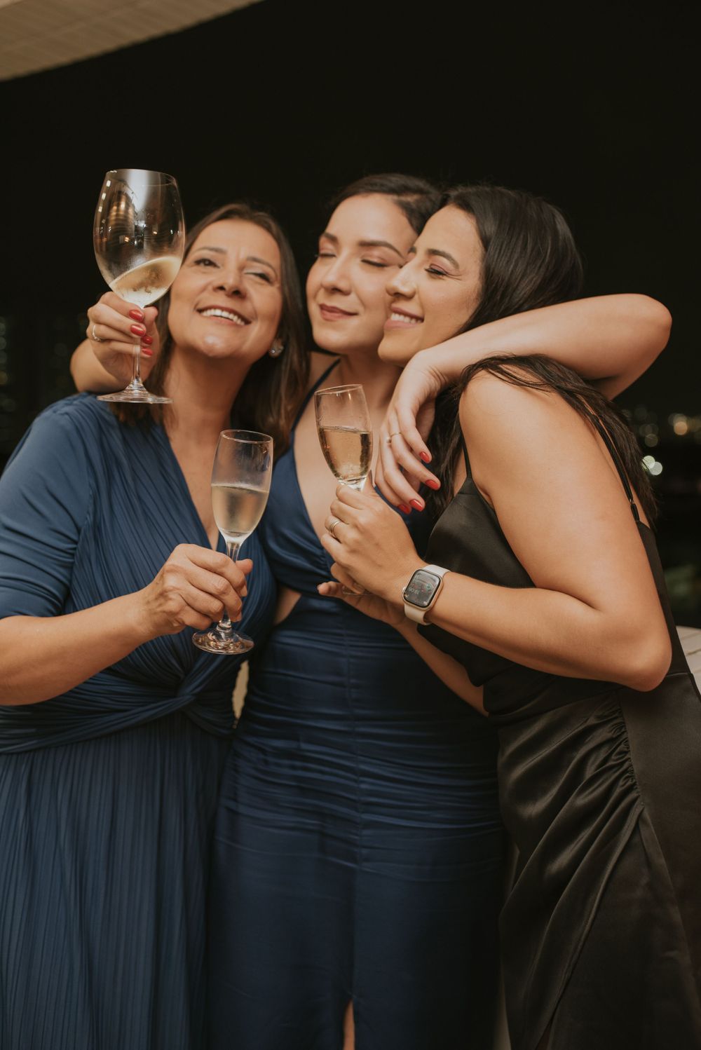 Three friends in elegant blue dresses share a joyful moment while toasting with champagne glasses at an evening event.