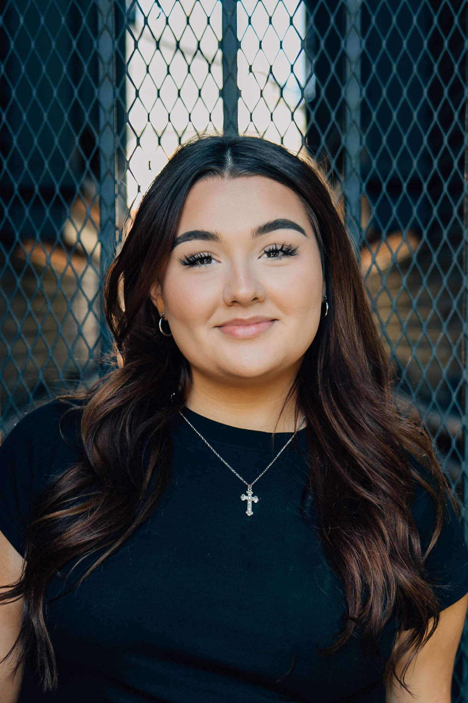 Young woman with long dark hair wearing a black top and cross necklace smiling at camera.