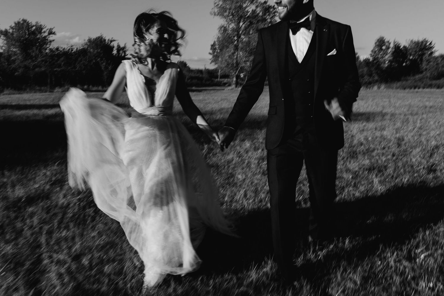 A black and white photo of newlyweds running playfully through a field.
