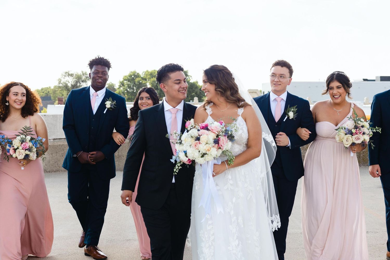 Wedding party walks together and smiles during outdoor celebration with pink bridesmaid dresses and white floral bouquets.