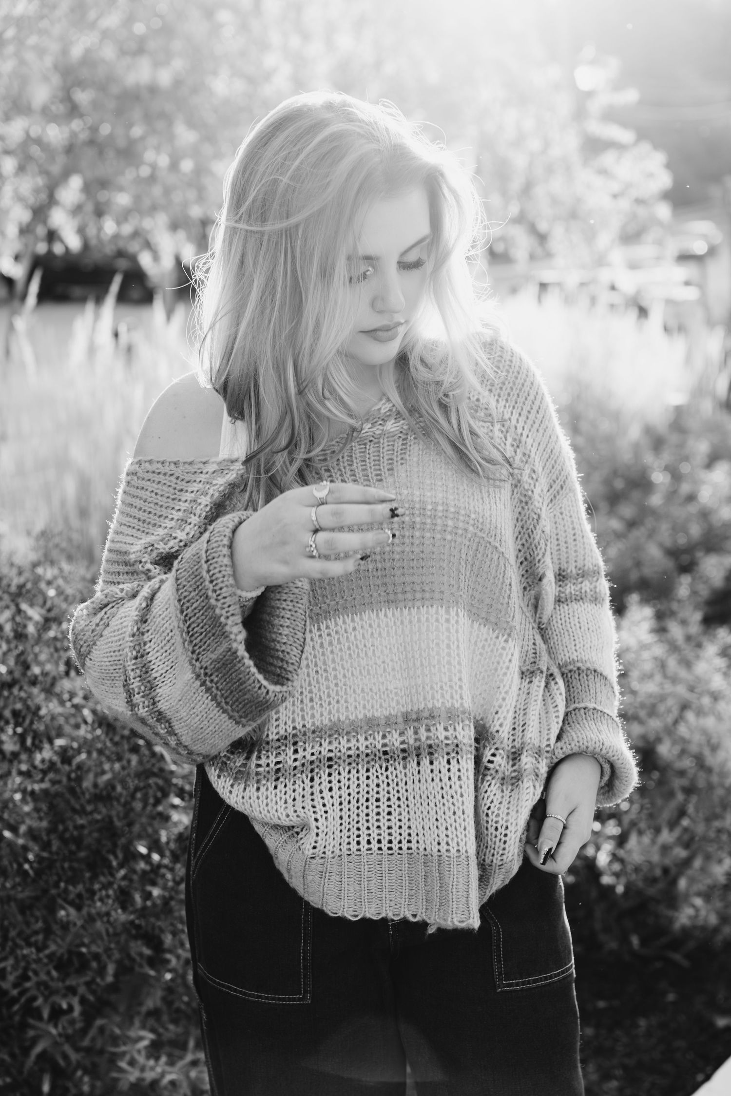 A black and white portrait showing someone in a chunky knit sweater looking down while standing in soft natural light outdoors.