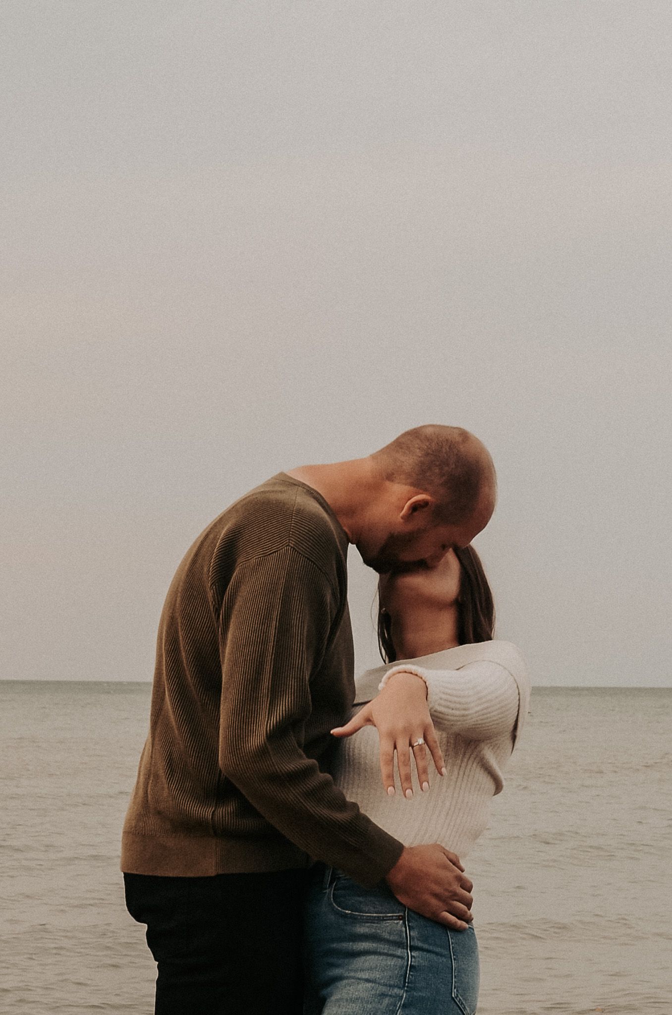 A romantic couple embraces beside cloudy waters on an overcast day.