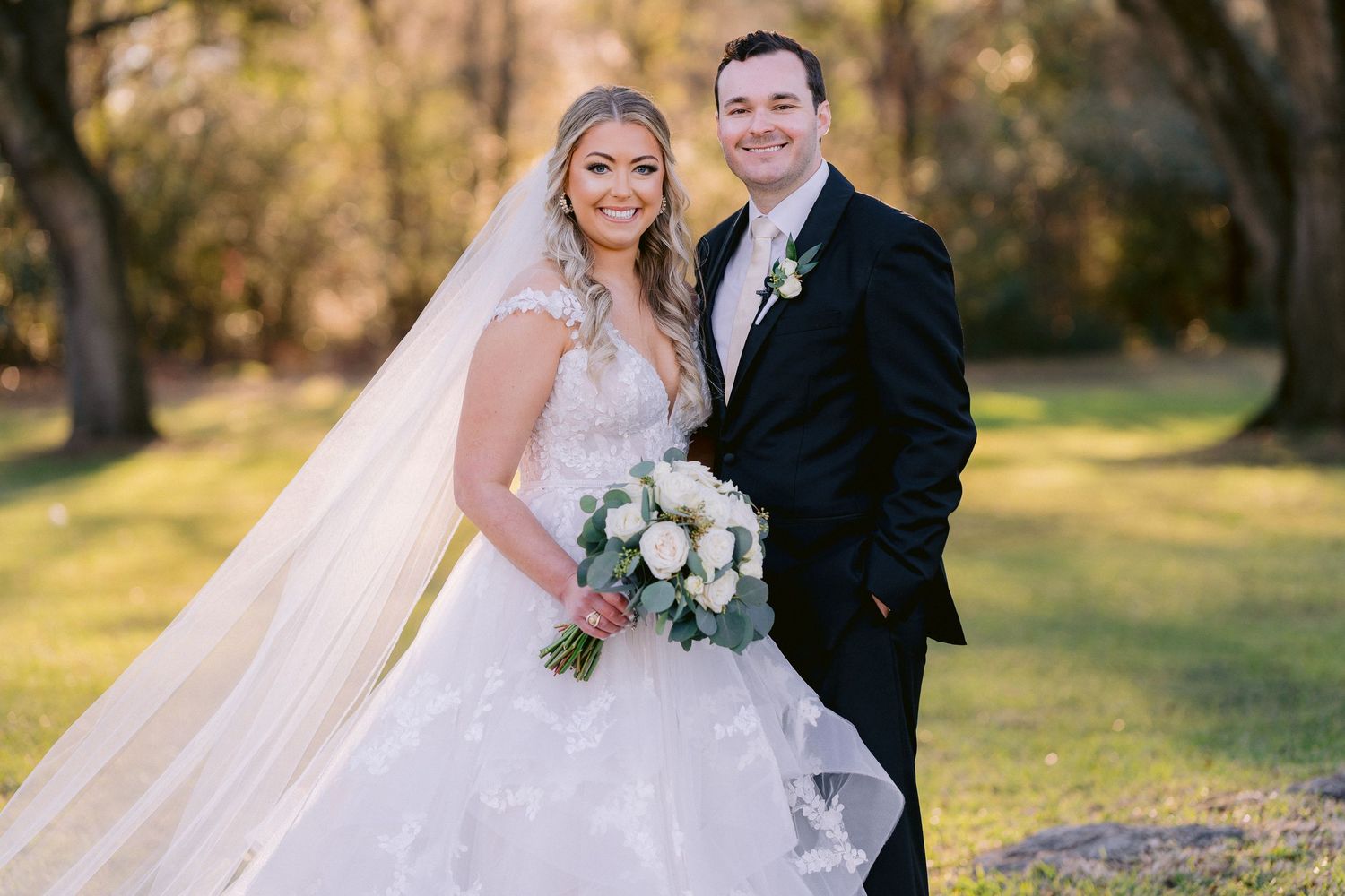 A bride in a lacy white dress with a long veil holds a white flower bouquet while posing outdoors with her groom.