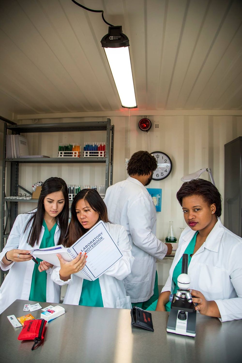 Medical students in white lab coats and green scrubs collaborate while working with microscopes and documents in a laboratory.