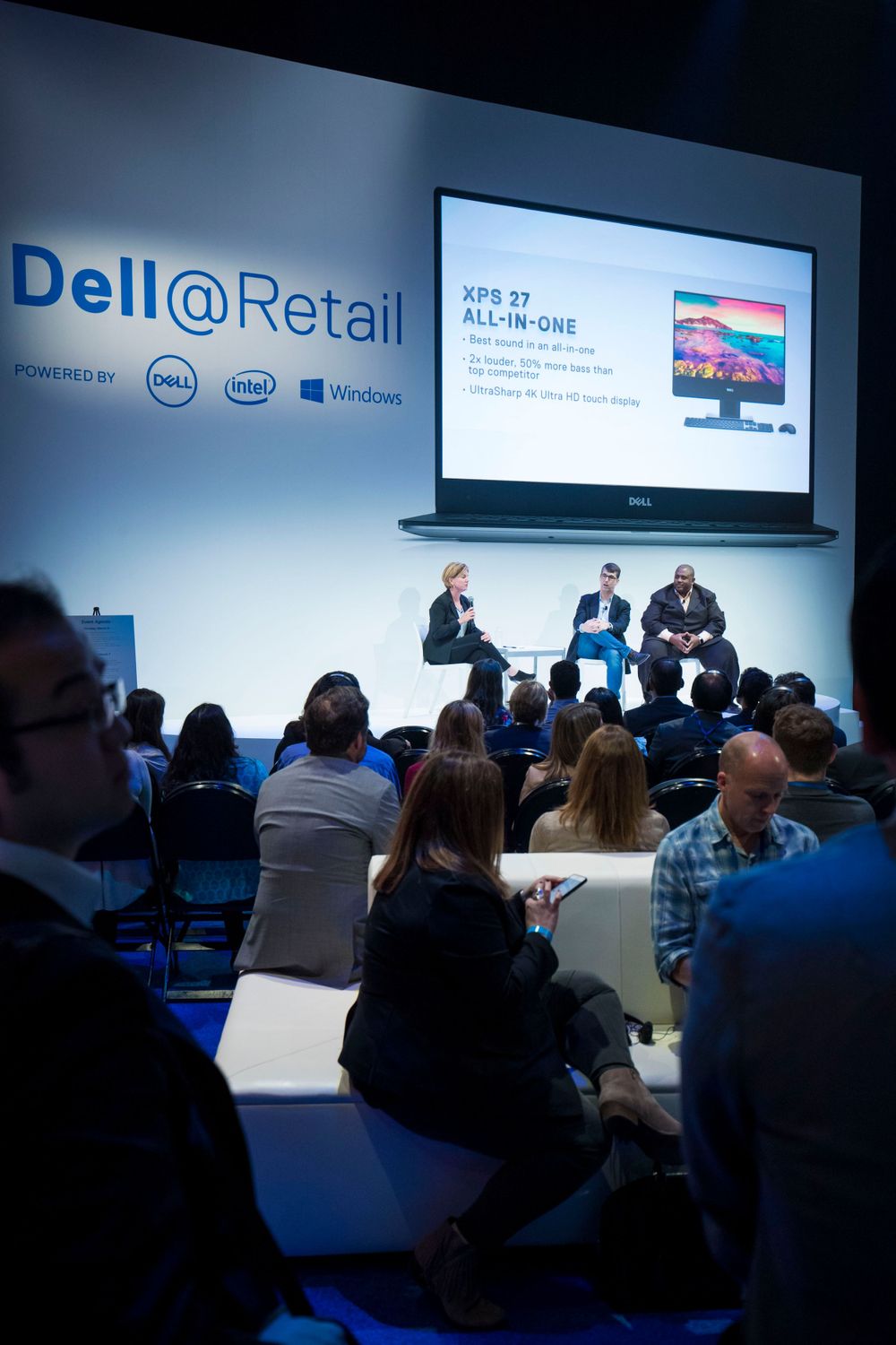 Dell retail presentation in conference room with audience seated in blue chairs and large projection screen display.