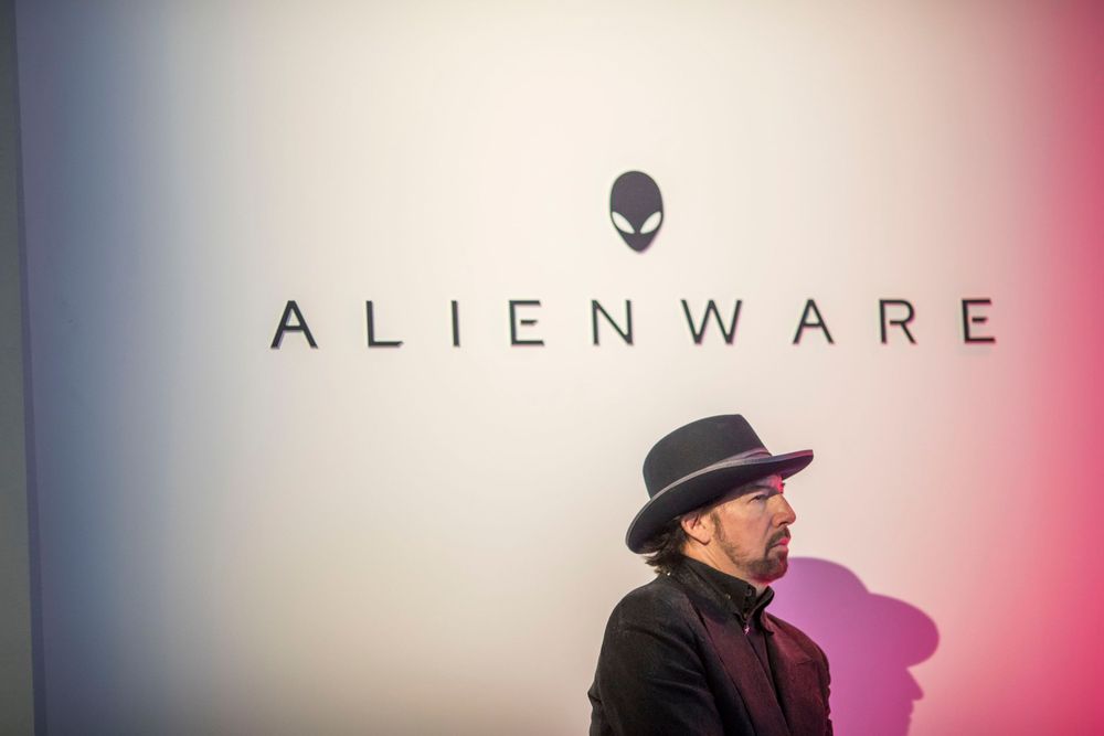 A person in a fedora stands in profile against a wall displaying the Alienware logo and company branding.