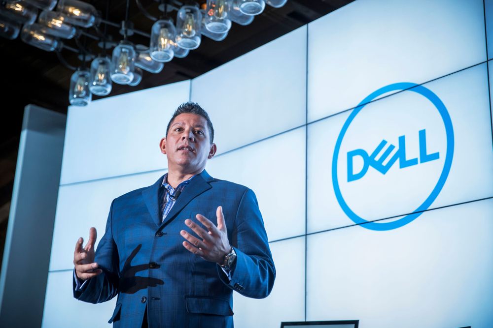 A presenter speaks on stage at a corporate event with the Dell logo displayed on a large screen behind them.
