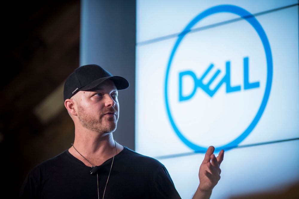 Someone wearing a black baseball cap and shirt stands in front of an illuminated Dell logo on a blue-lit wall.