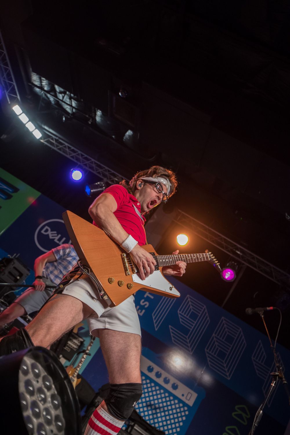 Musician in red shirt and white shorts plays electric guitar on stage with colorful lighting at a performance.