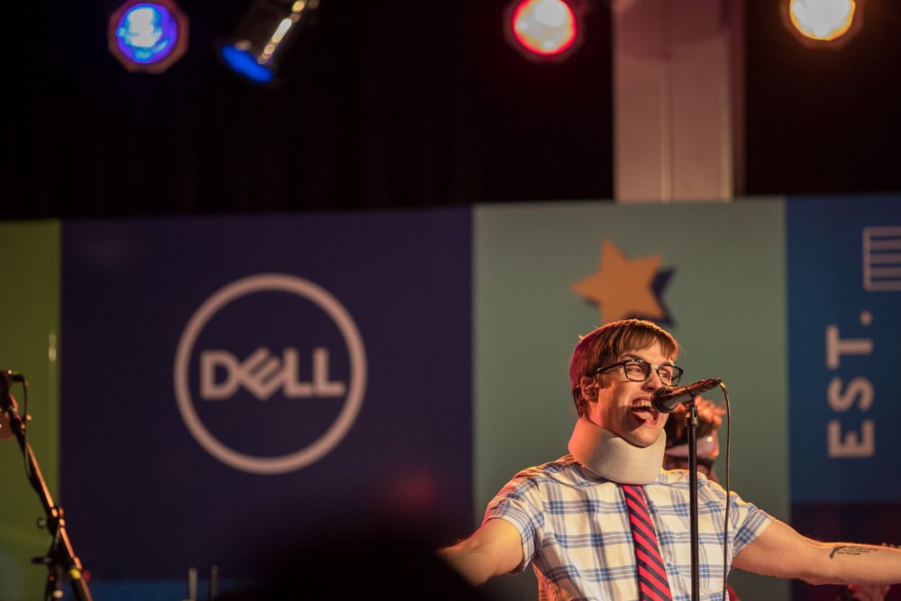 A performer sings into a microphone on stage with a Dell logo visible in the background lighting.