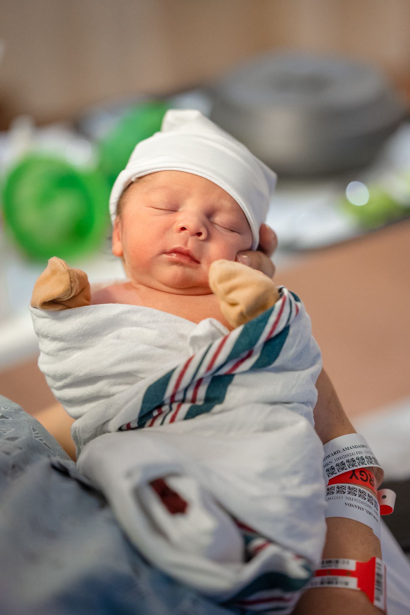 A peaceful newborn baby sleeps swaddled in a white and striped blanket wearing a small white cap in a hospital.