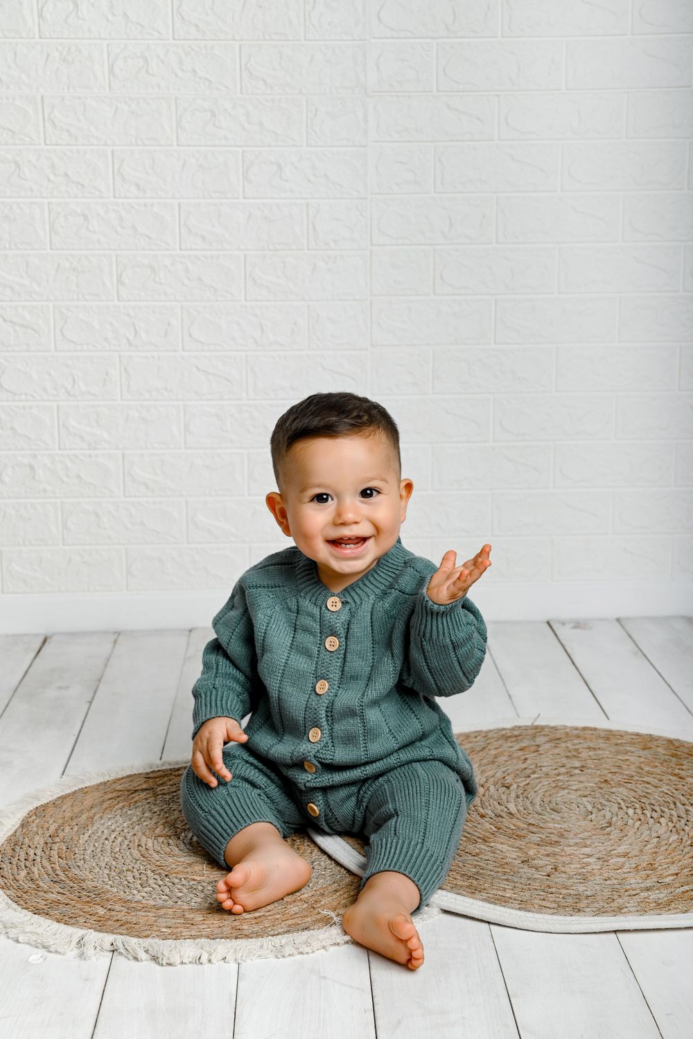 Smiling toddler wearing a sage green button-up linen outfit sits on round jute rug against white backdrop.