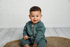 Sweet little one wearing a teal sweater smiles while sitting on a round woven rug against white backdrop.