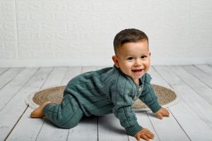 Baby wearing a green sweater crawls on a rustic wooden floor near a burlap mat.