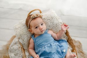 Baby in light blue dress lies on lace blanket in wicker basket during professional photo session.