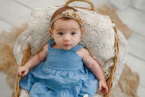 A small infant wearing a light blue dress lies in a woven basket lined with white lace fabric.