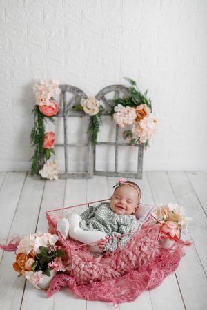 Sweet baby resting on pink lace blanket with vintage window frame and floral decorations in studio setting.