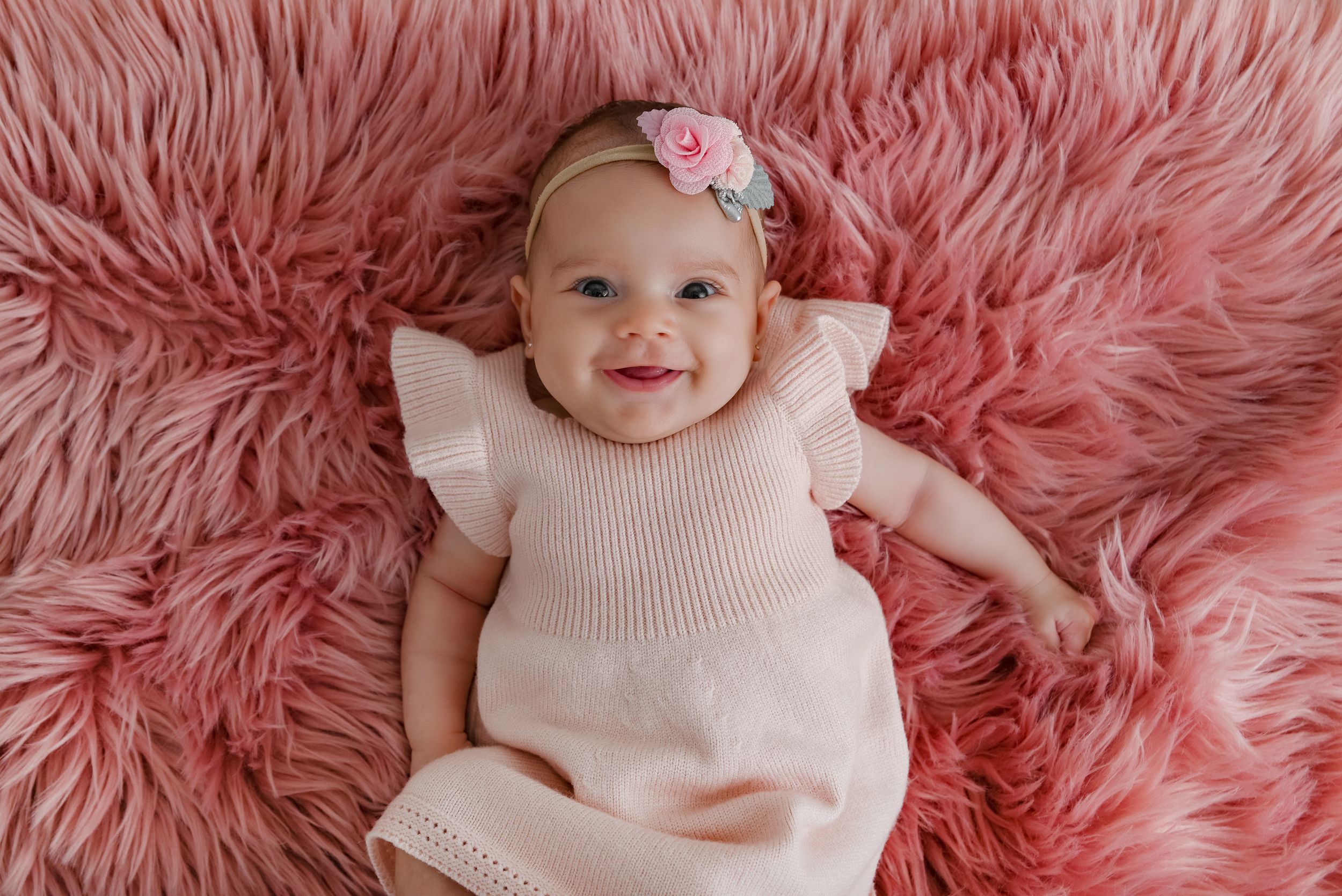 Baby in cream colored dress lying on pink fluffy blanket smiling with floral headband.