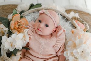 Baby wearing pink sweater and headband lies in wicker basket surrounded by peach and white floral decorations.