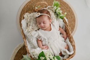 Sleeping newborn wearing white dress and headband in wicker basket decorated with delicate white flowers and greenery.