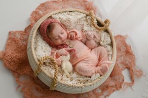 Sweet newborn baby sleeps peacefully in round basket surrounded by soft peach-colored fabric and delicate decorative elements.