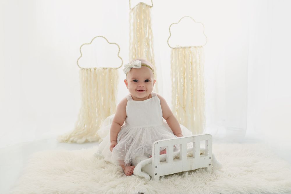 Baby in a white dress sitting beside a small decorative fence against a backdrop with painted clouds and trees.
