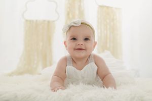 Small baby wearing white clothing lies on soft white fur blanket with light curtains in background.