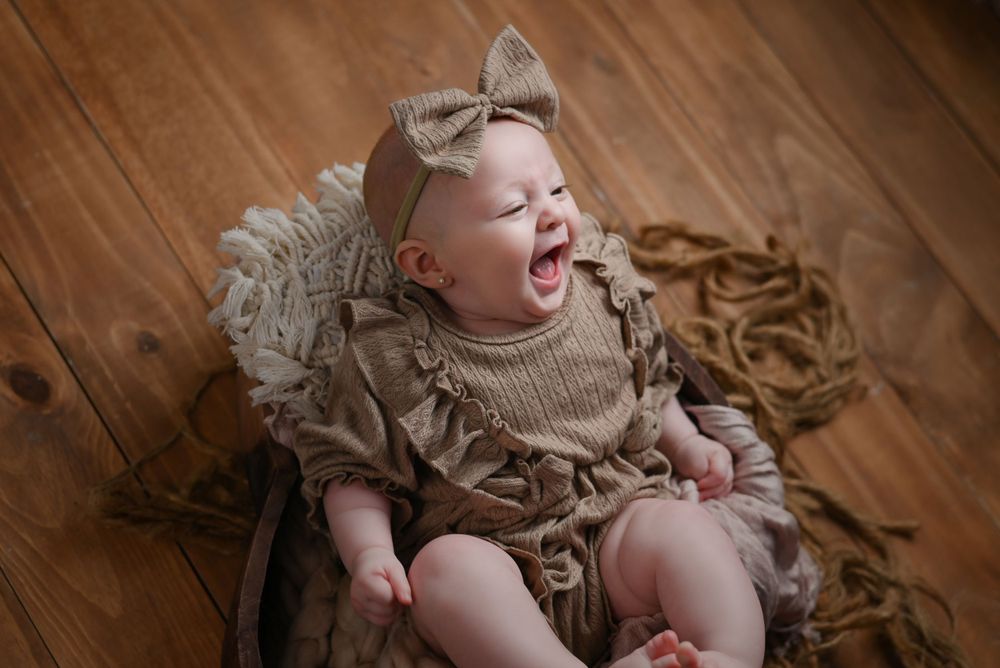 Baby wearing a beige bow and sweater sits on a wooden floor laughing joyfully during a photo session.