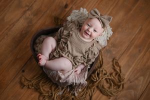 Smiling baby wearing ruffled beige dress lying on wooden floor surrounded by flowing fabric.