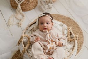 Baby in cream colored outfit rests on round woven mat with macrame decorations and soft natural decor.