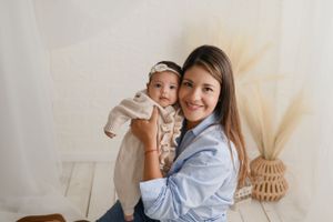 A woman in a blue button-down shirt holds a baby in a cream-colored outfit against a neutral backdrop with rattan decor.