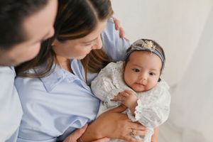 Baby wearing white lace dress and headband being held in loving embrace against light background.