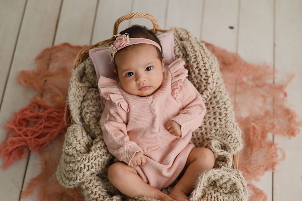 Baby wearing pink outfit lying on knitted blanket and soft peach-colored backdrop during photo session.