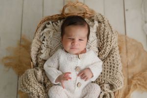A newborn baby rests peacefully in a cozy beige knit blanket against a white wooden backdrop.