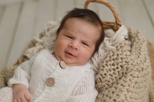 A peaceful sleeping baby wearing a white knitted sweater rests in a cozy basket with beige knit blankets.
