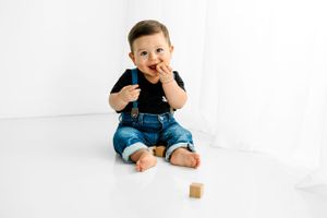 Small child sitting on white floor wearing blue jeans and black shirt with playful expression and wooden block nearby.