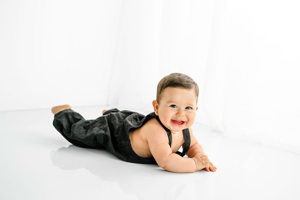Baby wearing dark pants lies on their stomach on a white backdrop smiling with a happy expression.