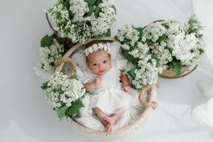 Baby in white dress lying in woven basket surrounded by white lilac flowers.
