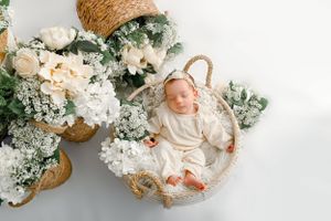 A baby rests peacefully in a woven basket surrounded by white floral arrangements and greenery against a light background.