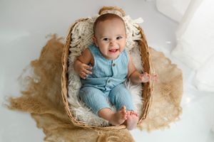 A baby wearing light blue overalls sits in a cozy basket lined with soft cream colored fabric and fur.