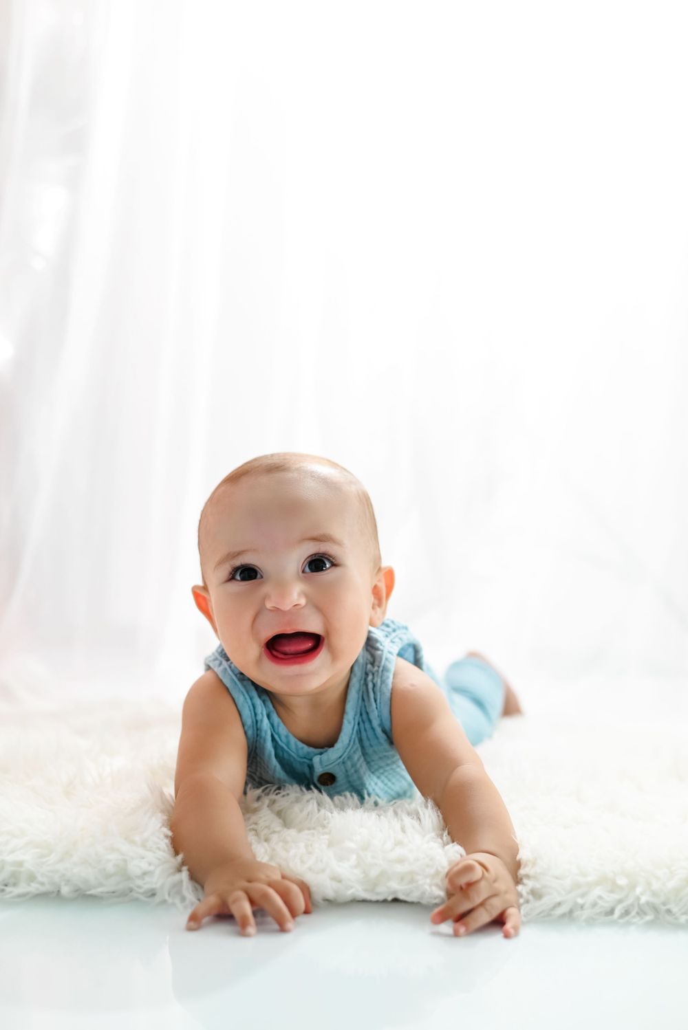 A baby wearing a blue sleeveless outfit lies on a white fluffy rug and smiles brightly.