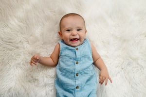 A happy baby wearing a light blue button-up romper lies on a white fluffy rug.