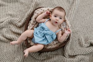 A baby wearing a light blue ruffled romper lies on a textured beige blanket in a cozy setting.